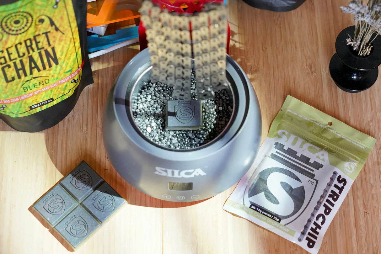 Silca Strip chip inside wax melting pot with SRAM Flattop chain hanging on the stand. Strip Chip packaging on the wooden counter next to it.